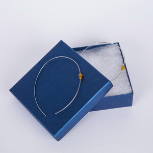 May include: A pair of silver-toned hoop earrings with small yellow accents, presented in an open, dark blue jewelry box lined with white cotton. The earrings have a minimalist design.