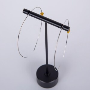 May include: A pair of silver-colored hoop earrings displayed on a black earring stand. The earrings have a simple, minimalist design with a curved shape. The stand is black with a round base and a horizontal bar to hold the earrings.