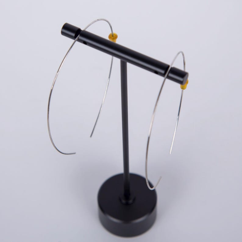 May include: A pair of silver-colored hoop earrings displayed on a black earring stand. The earrings have a simple, minimalist design with a curved shape. The stand is black with a round base and a horizontal bar to hold the earrings.