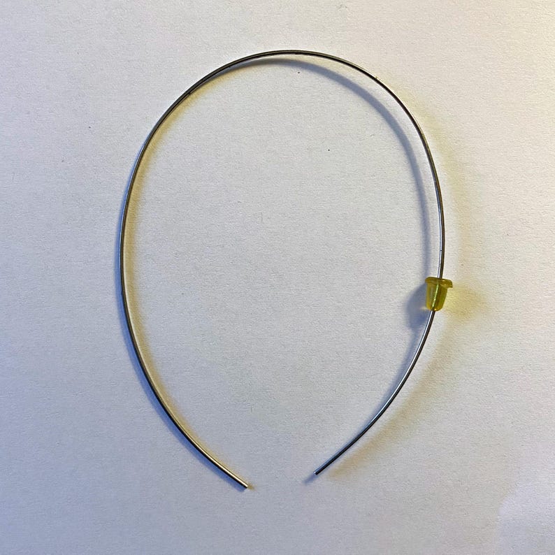 May include: A silver-colored metal headband with a small, yellow plastic piece attached. The headband is curved and open at the bottom. The background is a plain, light-colored surface.
