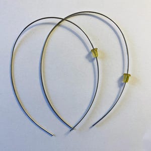 May include: A pair of silver-toned wire hoop earrings with yellow rubber earring backs. The earrings are curved and have a simple, minimalist design, set against a white background.
