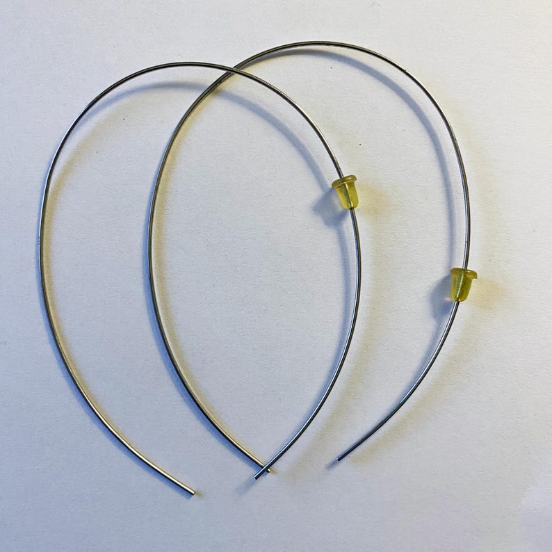 May include: A pair of silver-toned wire hoop earrings with yellow rubber earring backs. The earrings are curved and have a simple, minimalist design, set against a white background.