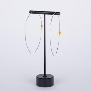 May include: A pair of silver-toned hoop earrings with yellow accents are displayed on a black earring stand. The earrings have a minimalist design, with a slender, curved shape. The stand has a round base and a vertical post.
