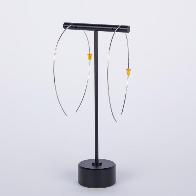 May include: A pair of silver-toned hoop earrings with yellow accents are displayed on a black earring stand. The earrings have a minimalist design, with a slender, curved shape. The stand has a round base and a vertical post.