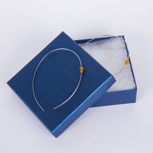 May include: A pair of silver-toned hoop earrings with small, amber-colored beads, presented in an open, textured, navy blue gift box with white padding. The earrings have a simple, elegant design.