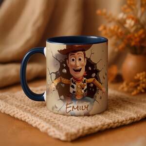 May include: A ceramic mug with a cartoon image of Woody from Toy Story breaking through a wall. The mug has a blue handle and interior. The name "Emily" is written on the front. The mug is on a beige knitted coaster.