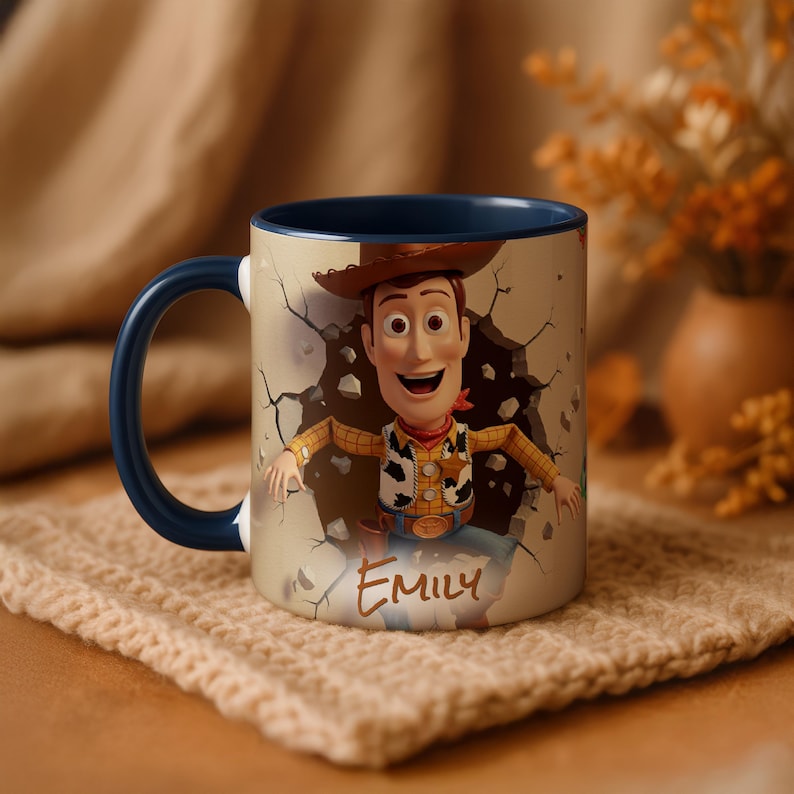 May include: A ceramic mug with a cartoon image of Woody from Toy Story breaking through a wall. The mug has a blue handle and interior. The name "Emily" is written on the front. The mug is on a beige knitted coaster.
