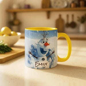 May include: A white ceramic mug with a yellow handle and interior. The mug features a cartoon image of Olaf from Frozen, with the name "Emily" printed below. The background shows a snowy scene.