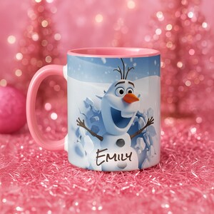 May include: A white ceramic mug with a pink handle and interior. The mug features a cartoon image of Olaf the snowman from Frozen, emerging from a snowy background. The name "Emily" is written on the mug in a script font.