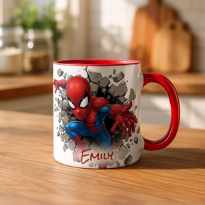 K&ouml;nnte beinhalten: Wei&szlig;er Keramikbecher mit Spider-Man, der durch eine Mauer aus grauem Stein bricht. Der Becher hat einen roten Innenraum und Henkel, mit dem Namen "Emily" in roter Schrift. Ein lustiges und farbenfrohes Geschenk f&uuml;r Superhelden-Fans.