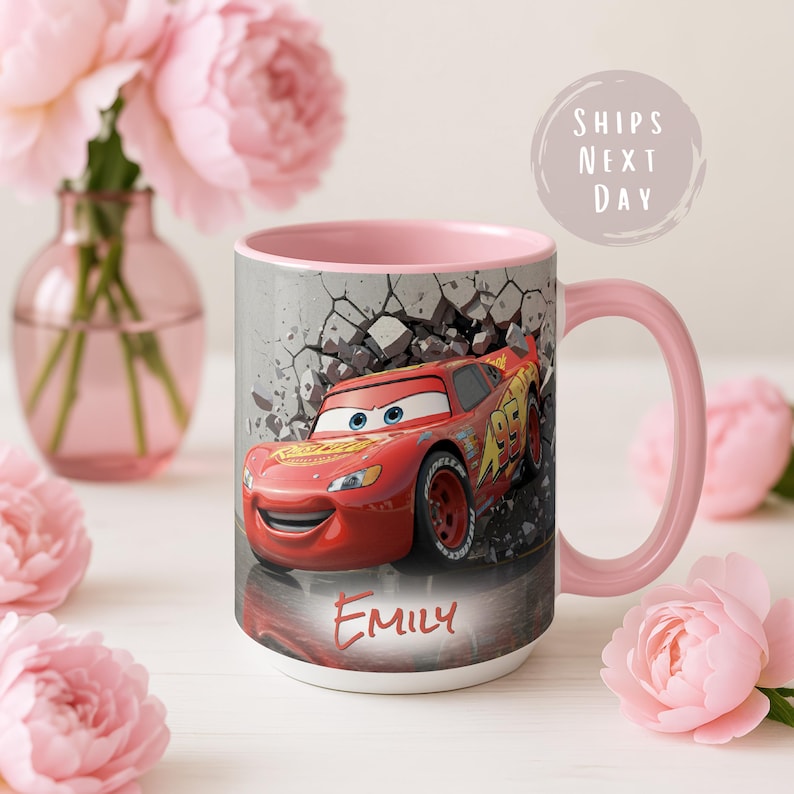 May include: A white ceramic mug with a pink interior and handle, featuring a red race car design. The car is breaking through a gray wall, with the name "Emily" printed below. The mug is surrounded by pink flowers.
