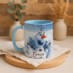 May include: A ceramic mug with a light blue handle and interior. The mug features a smiling Olaf from Frozen, with the name "Emily" printed below. The background shows a snowy scene. The mug is on a wooden coaster.