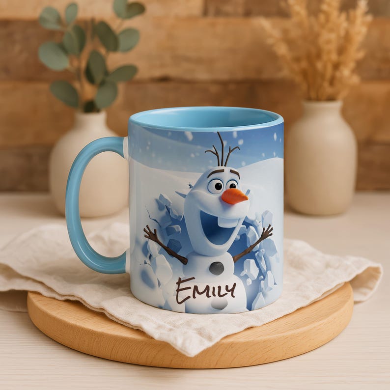 May include: A ceramic mug with a light blue handle and interior. The mug features a smiling Olaf from Frozen, with the name "Emily" printed below. The background shows a snowy scene. The mug is on a wooden coaster.
