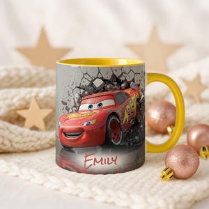 May include: A ceramic mug with a yellow handle and interior. The exterior features a red race car graphic, with the name "Emily" below. The background shows a gray wall with a cracked design.