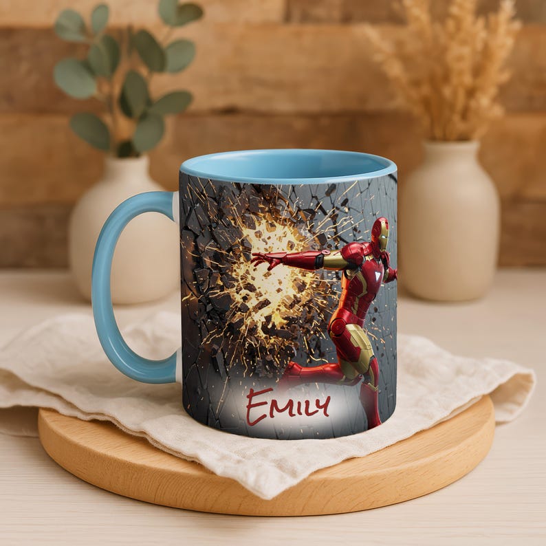 May include: A ceramic mug with a light blue interior and handle. The exterior features a graphic of Iron Man breaking through a wall, with the name "Emily" in red script. The mug is on a wooden coaster.