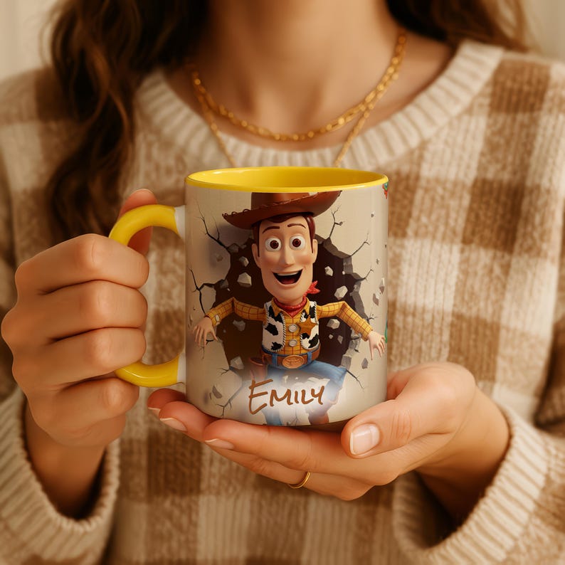 May include: A ceramic mug with a cartoon image of Woody from Toy Story, bursting through a wall. The mug has a yellow handle and interior, with the name "EMILY" printed below the character. The mug is held by a person.