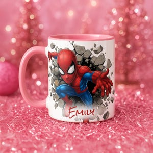 K&ouml;nnte beinhalten: Wei&szlig;er Keramikbecher mit rosa Griff und Rand, mit einer Grafik von Spider-Man, der durch eine Wand bricht. Der Name "Emily" ist in roter Schrift auf der Vorderseite geschrieben. Der Hintergrund ist eine rosa, glitzernde Oberfl&auml;che.