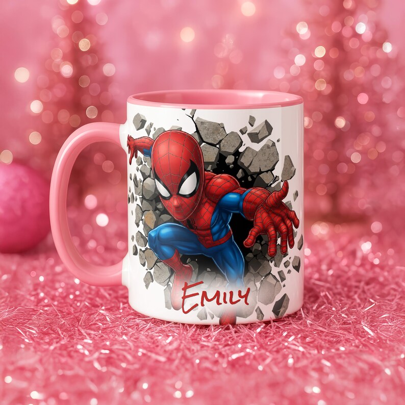 K&ouml;nnte beinhalten: Wei&szlig;er Keramikbecher mit rosa Griff und Rand, mit einer Grafik von Spider-Man, der durch eine Wand bricht. Der Name "Emily" ist in roter Schrift auf der Vorderseite geschrieben. Der Hintergrund ist eine rosa, glitzernde Oberfl&auml;che.