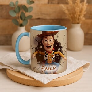 May include: A ceramic mug with a cartoon character, Woody, breaking through a wall design. The mug has a light blue interior and handle. The name "Emily" is printed on the front. The mug is on a wooden coaster with a white cloth.