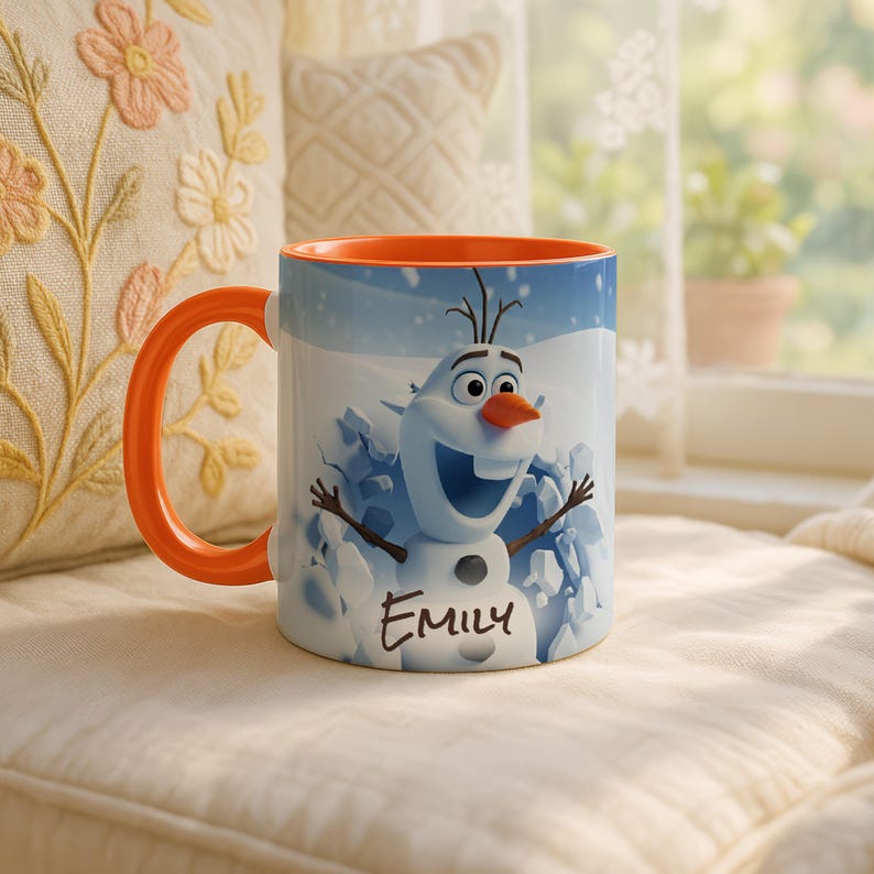 May include: A white ceramic mug featuring Olaf from Frozen, with an orange handle and interior. The mug has a snowy background and the name "Emily" printed on it. The mug is sitting on a white cushion.