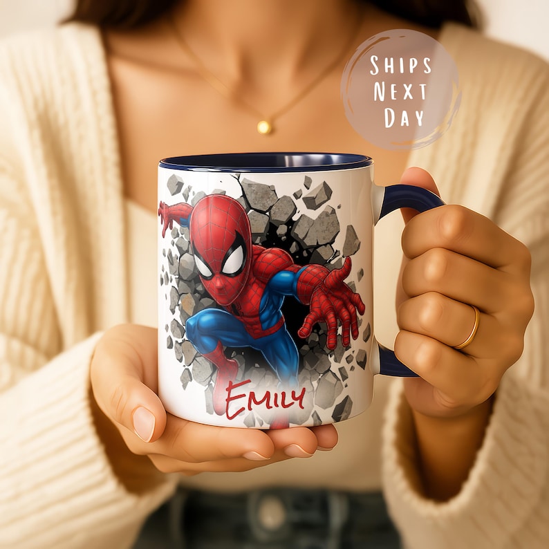 K&ouml;nnte beinhalten: Wei&szlig;er Keramikbecher mit blauem Innenraum und Henkel, der Spider-Man zeigt, der durch eine Wand bricht. Auf dem Becher steht der Name "Emily" in Rot. Der Text "Ships Next Day" ist sichtbar. Der Becher wird von einer Person gehalten.
