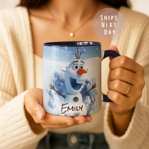 May include: A blue and white ceramic mug featuring Olaf from Frozen, with the name "Emily" printed on the front. The mug has a dark blue handle and interior. The background shows a snowy scene.