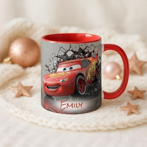 May include: A red and white ceramic mug featuring Lightning McQueen from the movie Cars. The mug has a red interior and handle, with the name "Emily" printed on the front. The background shows a cracked wall.