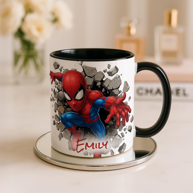 K&ouml;nnte beinhalten: Wei&szlig;er Keramikbecher mit Spider-Man, der durch eine Wand bricht. Der Becher hat einen schwarzen Innenraum und Henkel, und der Name "Emily" ist rot geschrieben. Der Becher steht auf einem silbernen Untersetzer.