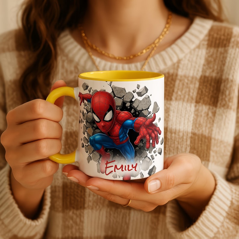 K&ouml;nnte beinhalten: Wei&szlig;e Keramiktasse mit gelbem Henkel und Innenraum. Die Tasse zeigt eine farbenfrohe Illustration von Spider-Man, der durch eine Wand bricht, mit dem Namen "Emily" in roter Schrift. Die Tasse wird von einer Person gehalten.