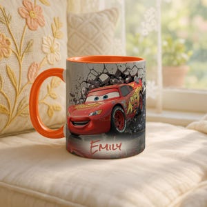 May include: A ceramic mug with an orange handle and interior. The exterior features a graphic of the character Lightning McQueen from the movie Cars, breaking through a wall. The name "Emily" is printed below the graphic.
