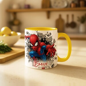 K&ouml;nnte beinhalten: Wei&szlig;er Keramikbecher mit gelbem Innenraum und Henkel. Der Becher zeigt eine Grafik von Spider-Man, der durch eine Wand bricht, mit dem Namen "Emily" in roter Schrift. Der Becher steht auf einer hellen Oberfl&auml;che.