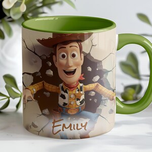 May include: A ceramic mug featuring a cartoon image of Woody from Toy Story breaking through a wall. The mug has a green handle and interior, with the name "Emily" printed on the front.