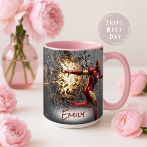 May include: A white ceramic mug with a pink interior and handle. The mug features a graphic of Iron Man breaking through a wall, with the name "Emily" below. The mug is surrounded by pink flowers.