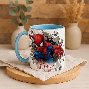 K&ouml;nnte beinhalten: Wei&szlig;er Keramikbecher mit hellblauem Innenraum und Henkel. Der Becher zeigt eine farbenfrohe Illustration von Spider-Man, der durch eine Wand bricht, mit dem Namen "Emily" in roter Schrift. Der Becher steht auf einem Holzuntersetzer.