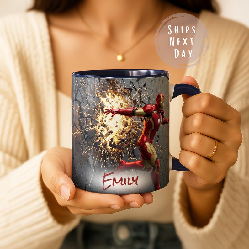 May include: A ceramic mug featuring a red and gold Iron Man design with a blue handle and interior. The mug has the name "Emily" printed on it. The mug has a design of Iron Man breaking through a wall. The mug is held by a person.