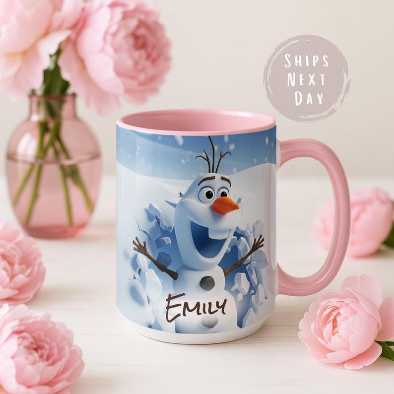 May include: A white ceramic mug featuring Olaf from Frozen, with a pink handle and interior. The mug has the name "Emily" printed on it. The background shows a winter scene with snow. The mug is surrounded by pink flowers.