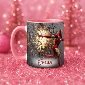 May include: A pink and white ceramic mug featuring Iron Man breaking through a wall. The mug has a pink handle and interior. The name "Emily" is written in red script on the front of the mug.
