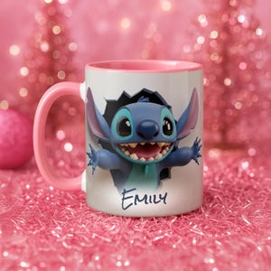Puede incluir: Una taza de cer&aacute;mica blanca con asa e interior rosa. La taza presenta un personaje de dibujos animados azul rompiendo una pared. El nombre "Emily" est&aacute; escrito en gui&oacute;n azul en la taza.