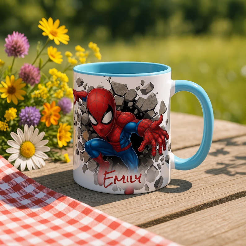 K&ouml;nnte beinhalten: Wei&szlig;er Keramikbecher mit blauem Innenraum und Henkel. Der Becher zeigt eine farbenfrohe Illustration von Spider-Man, der durch eine Wand bricht. Der Name "EMILY" steht in roter Schrift darunter. Der Becher steht auf einer Holzoberfl&auml;che.