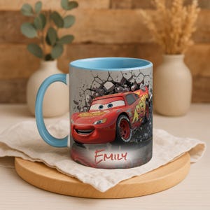 May include: A ceramic mug with a light blue interior and handle. The exterior features a red race car graphic with the name "EMILY" in red. The background shows a cracked wall.
