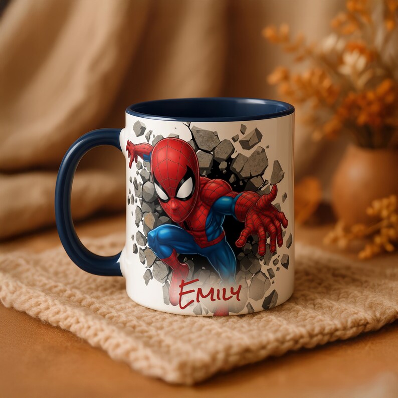 K&ouml;nnte beinhalten: Wei&szlig;er Keramikbecher mit blauem Henkel und Innenraum, der Spider-Man zeigt, der durch eine Wand bricht. Das Design umfasst Rot-, Blau- und Graut&ouml;ne, mit dem Namen "EMILY" in Rot. Ein farbenfroher Becher.