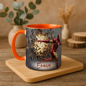 May include: Orange-handled mug featuring Iron Man breaking through a wall, with the name "Emily" in red script. The interior of the mug is orange, and it sits on a wooden tray.