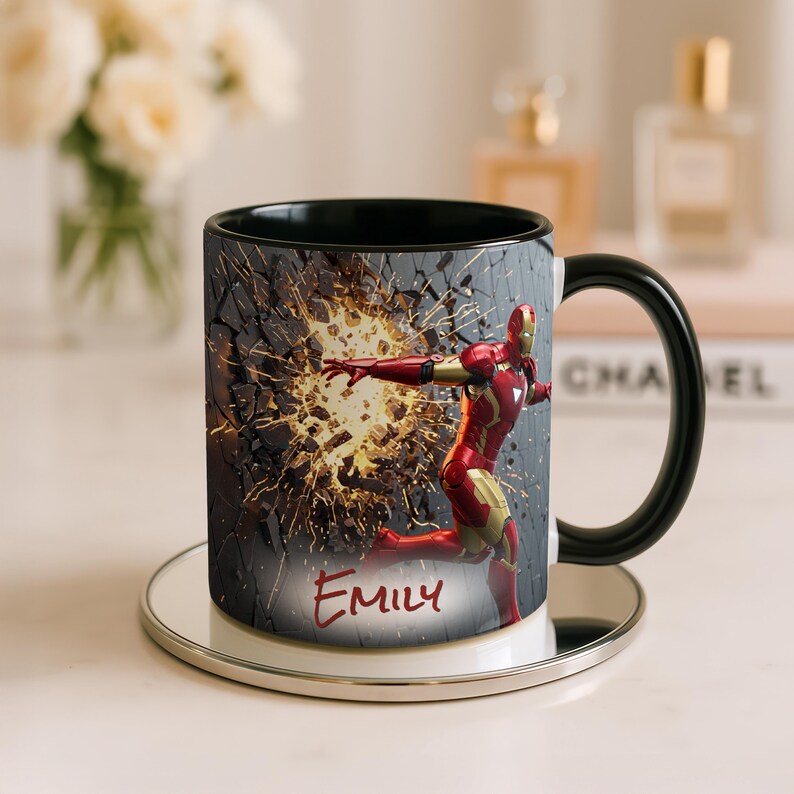 May include: Black and white ceramic mug featuring Iron Man in a dynamic pose, breaking through a wall. The mug has a black interior and handle, with the name "Emily" in red script. The mug sits on a silver coaster.