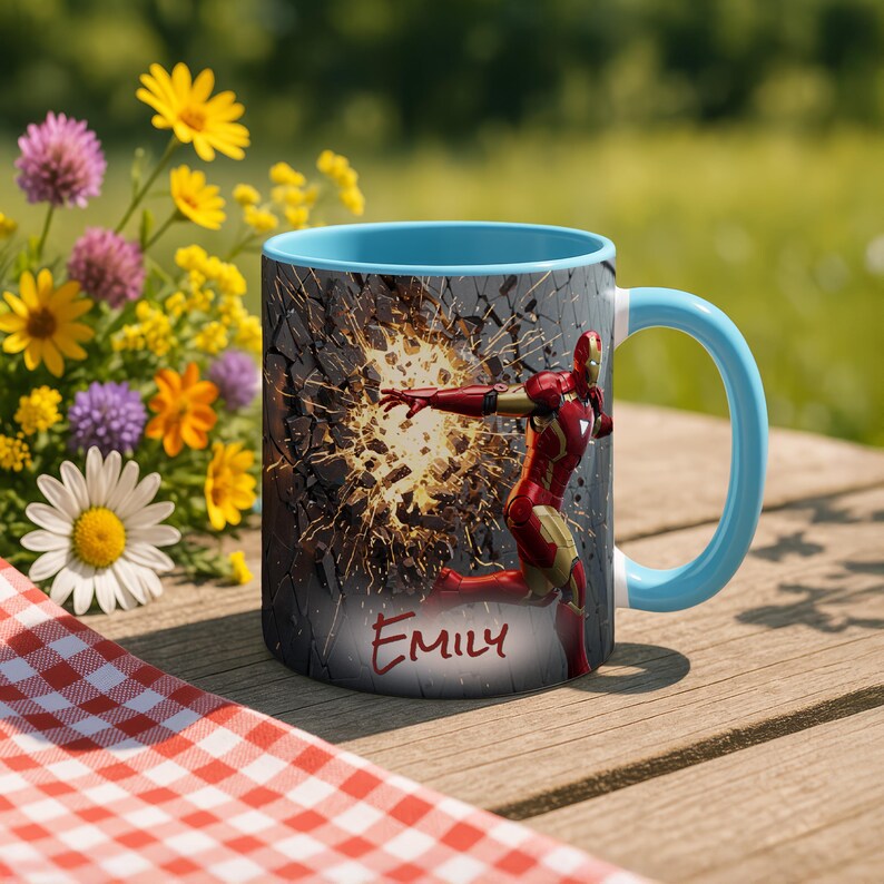 May include: A ceramic mug with a light blue interior and handle, featuring a red and gold Iron Man graphic breaking through a gray wall. The name "Emily" is written in red script below the graphic. The mug is on a wooden table with flowers.