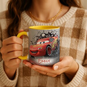 May include: A ceramic mug featuring Lightning McQueen from the Cars movie. The mug has a yellow handle and interior, with a design of the red race car breaking through a wall. The name "Emily" is written on the mug.
