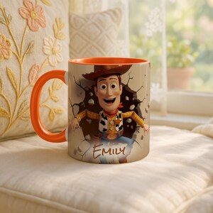 May include: A ceramic mug with a cartoon cowboy design, breaking through a wall. The mug has an orange handle and interior, with the name "Emily" printed on the front. The background includes a window and a decorative pillow.