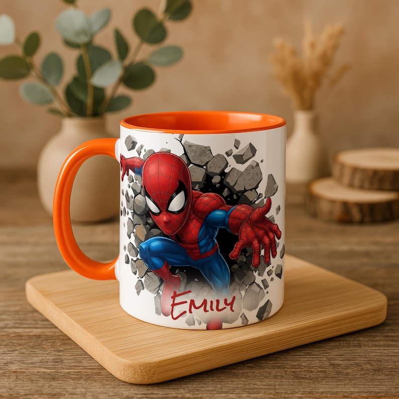 K&ouml;nnte beinhalten: Wei&szlig;e Keramiktasse mit orangefarbenem Innenraum und Griff. Die Tasse zeigt eine farbenfrohe Illustration von Spider-Man, der durch eine Wand bricht, mit dem Namen "EMILY" in roter Schrift. Die Tasse steht auf einem Holztablett.