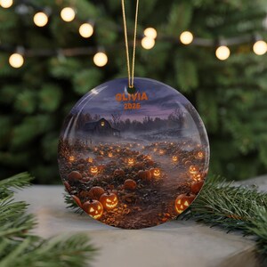 May include: A round ceramic ornament featuring a nighttime pumpkin patch scene with glowing jack-o'-lanterns, a rustic barn, and a misty landscape. The text OLIVIA 2026 is printed in orange at the top. The ornament hangs from a gold string.
