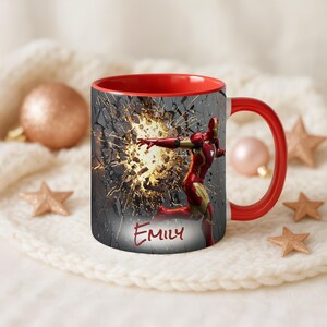 May include: A red and white ceramic mug featuring an Iron Man design. The mug's interior and handle are red. The Iron Man graphic shows the character breaking through a wall with the name "Emily" below.