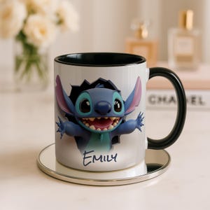 Puede incluir: Taza de cer&aacute;mica blanca con un personaje de dibujos animados azul, con orejas grandes y una amplia sonrisa, saliendo de un agujero dentado. La taza tiene un asa e interior negros, y el nombre "Emily" est&aacute; escrito debajo.